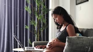 A beautiful pregnant Asian woman touching belly while working on laptop at home.